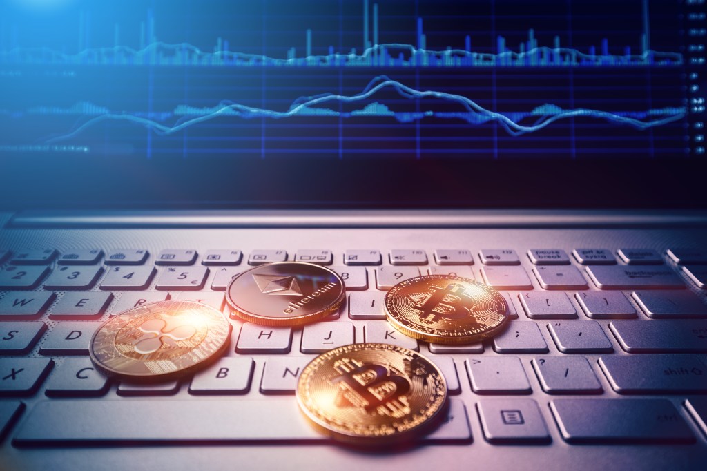 Four gold crypto coins, two Bitcoins, one Ethereum, and one Ripple, rest on a laptop keyboard with a blue financial chart on the screen.