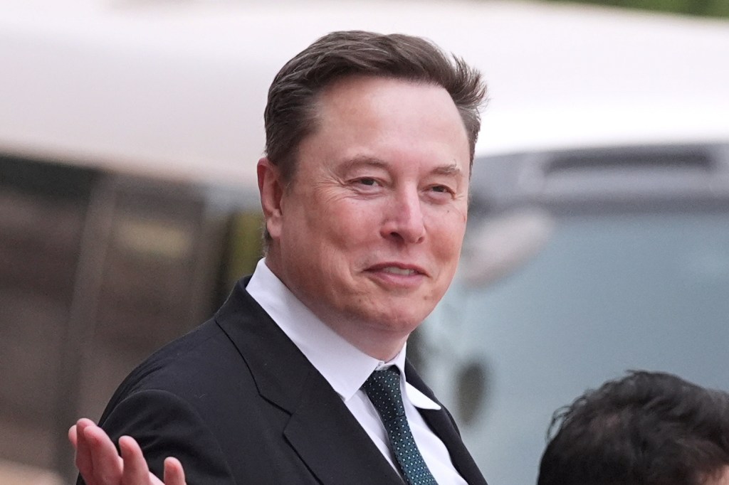 Elon Musk smiling and gesturing with his right hand while wearing a suit and tie.