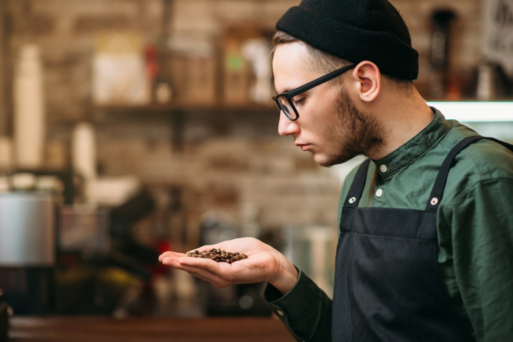A barista in a black beanie and apron holding a handful of coffee beans, smelling them.