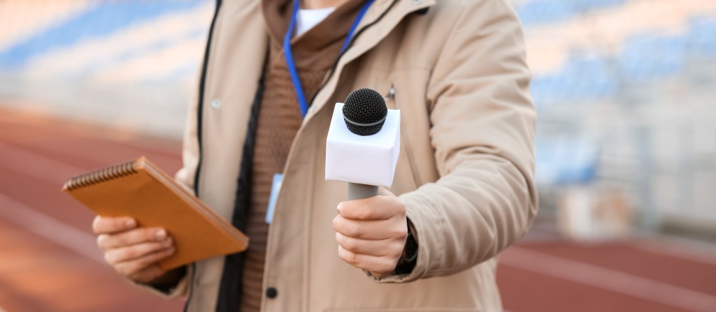 A male reporter holds a microphone and notepad at a stadium.