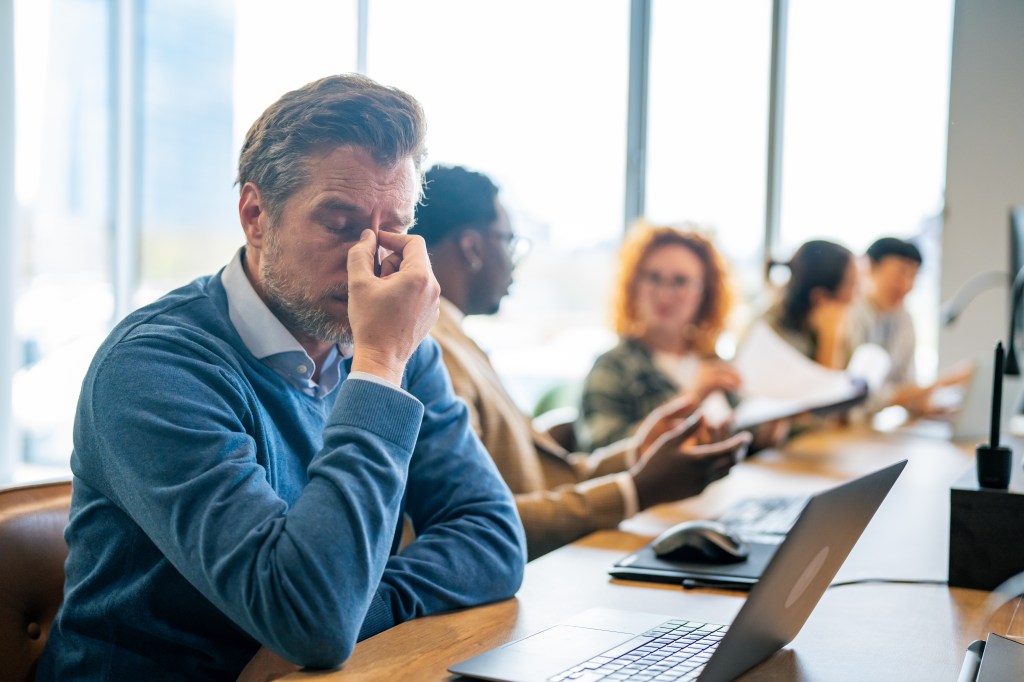 A stressed businessman with a headache rubs his eyes while working at a desk with blurred colleagues in the background.
