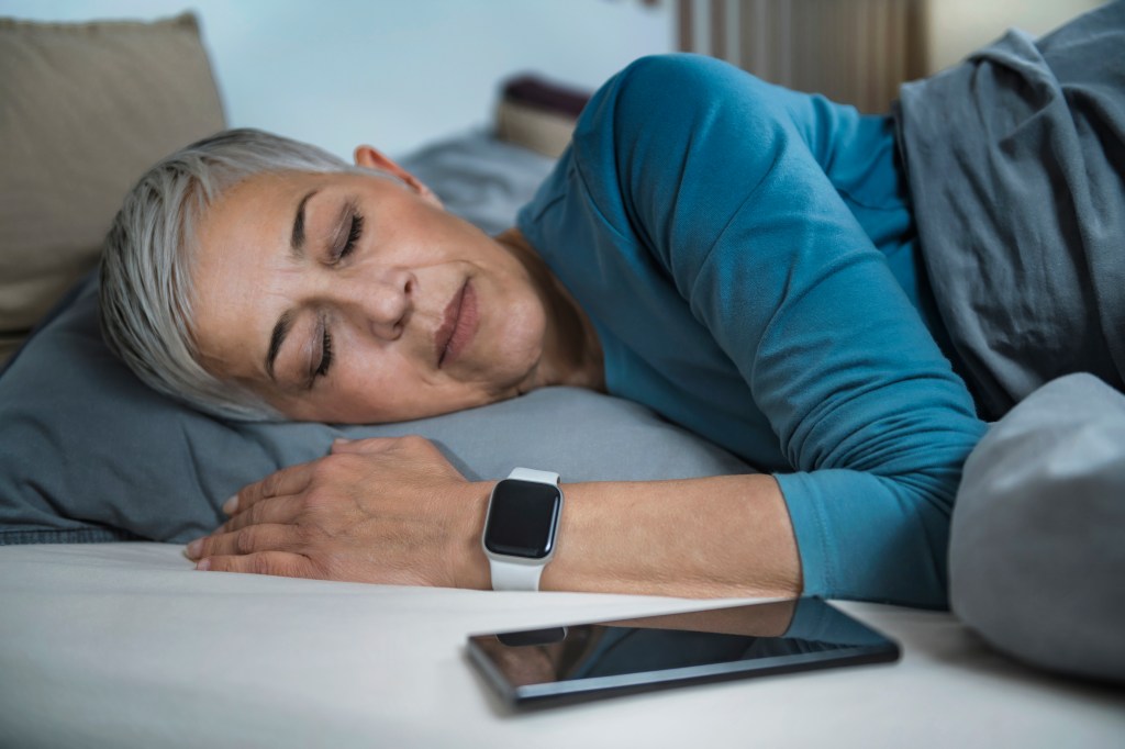 A senior woman sleeps in bed, using a smartwatch to track her sleeping habits, with a smartphone nearby.