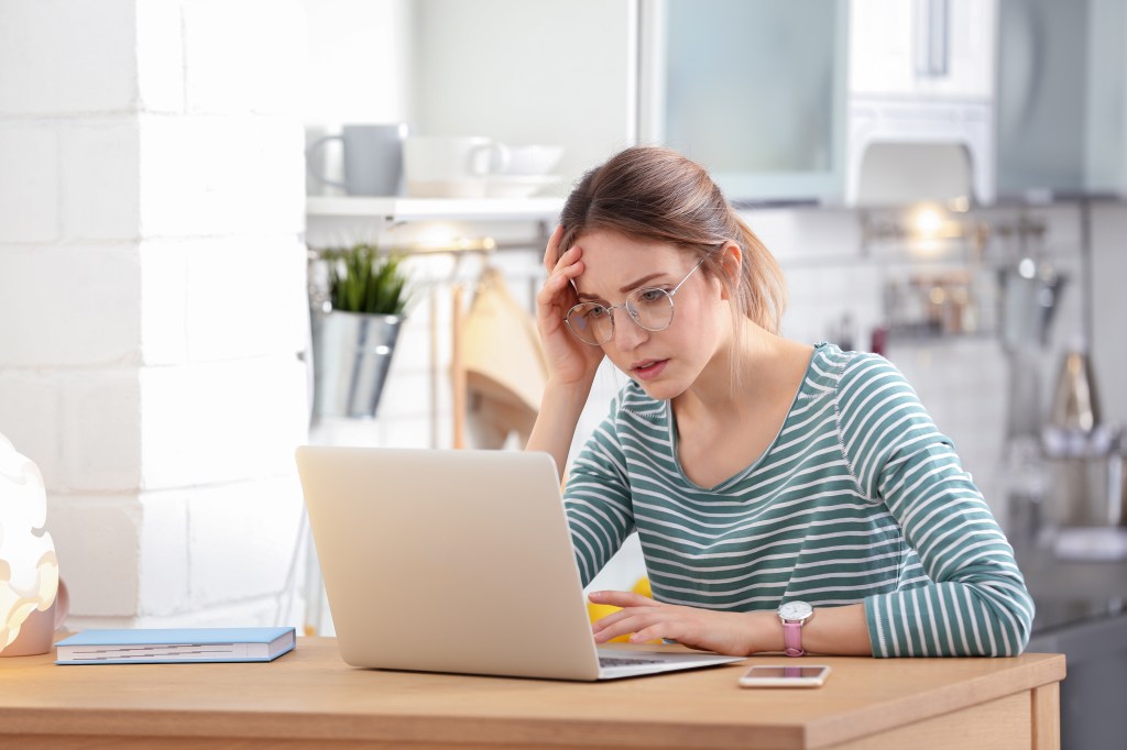 Troubled young woman in glasses and striped shirt working on a laptop at home.