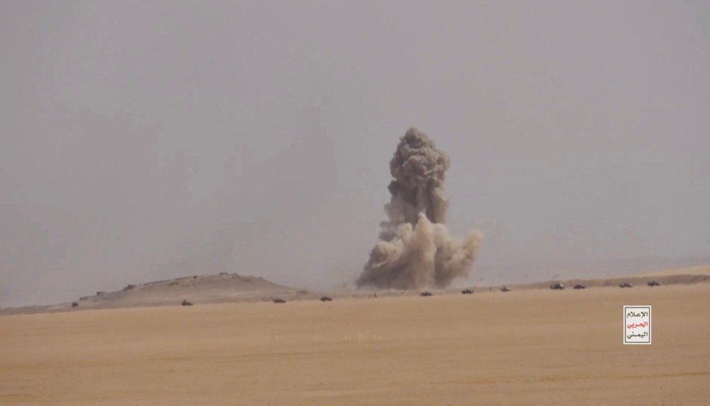 Smoke from an explosion billows into the sky above a desert landscape with military vehicles, depicting a battle between Iran-aligned Houthi fighters and Saudi-backed forces in al Jawf governate.