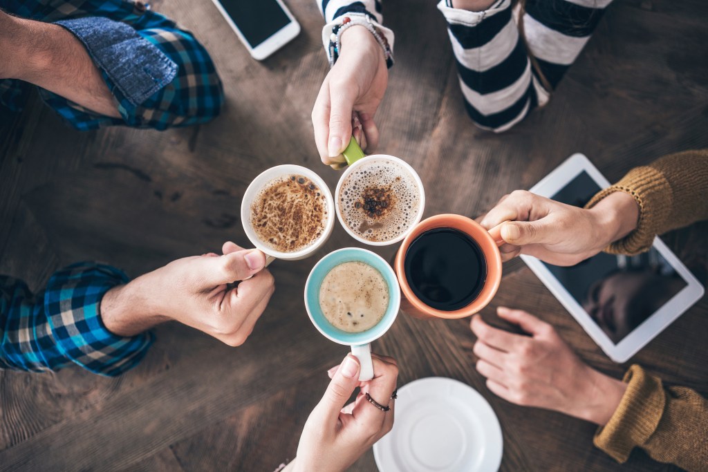 High-angle view of four people toasting coffee mugs together around a wooden table.