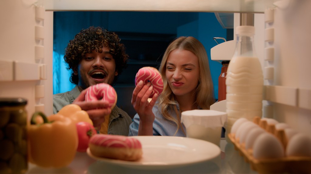 POV shot from inside a refrigerator of a couple eating donuts at night.