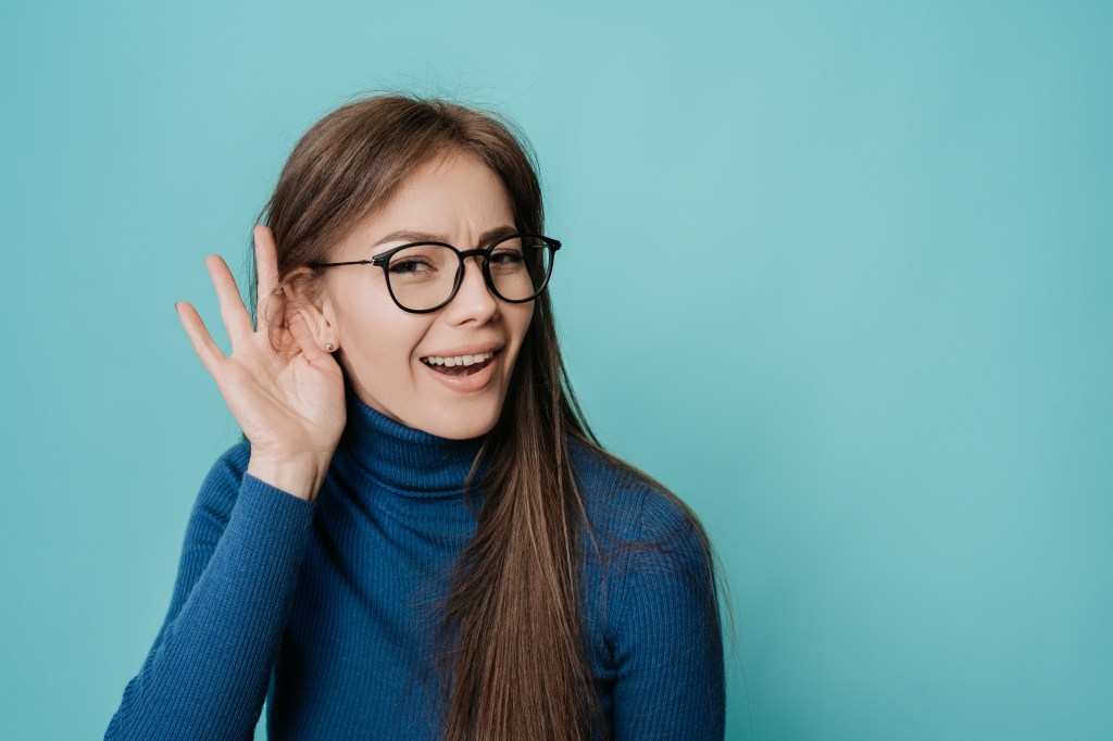 A young woman in glasses holding her hand to her ear, straining to listen.