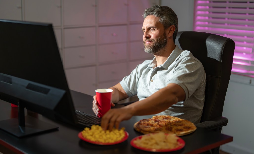 A man eats pizza and chips while drinking from a red cup at his computer.
