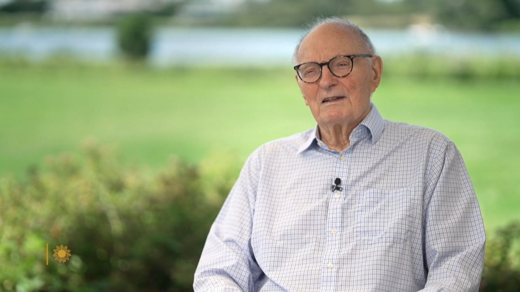 Alan Alda wearing glasses and a light-colored button-down shirt outdoors.