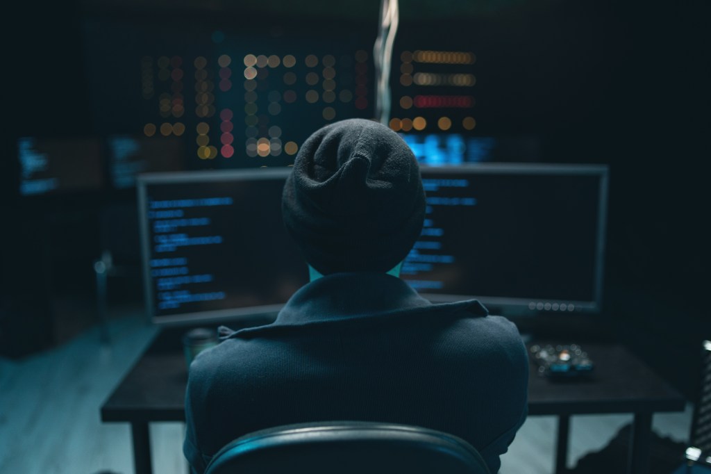 A hooded person in a dark room sits in front of computer monitors displaying code.