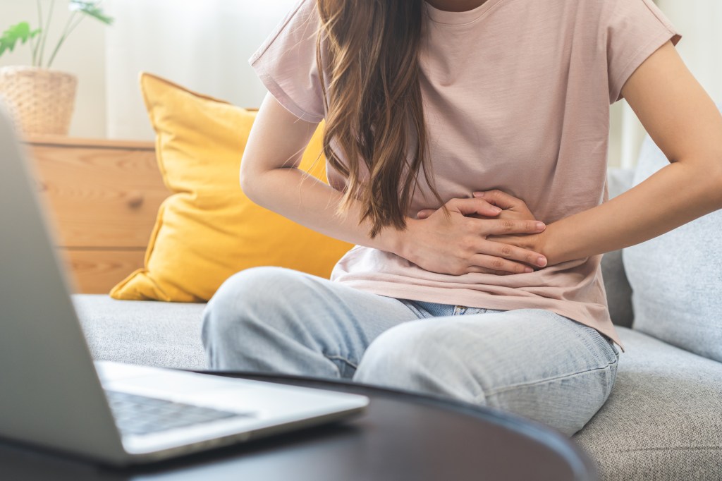 An Asian woman holding her stomach in pain while sitting on a couch in front of a laptop.