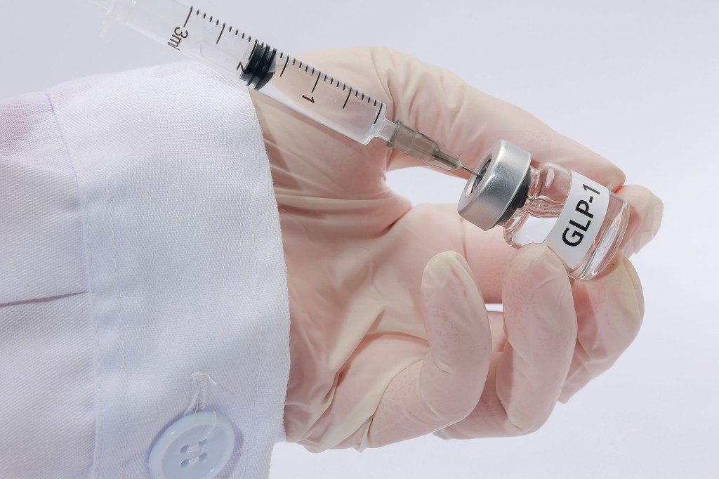 A gloved hand in a white lab coat prepares a GLP-1 injection from a labeled vial using a syringe.