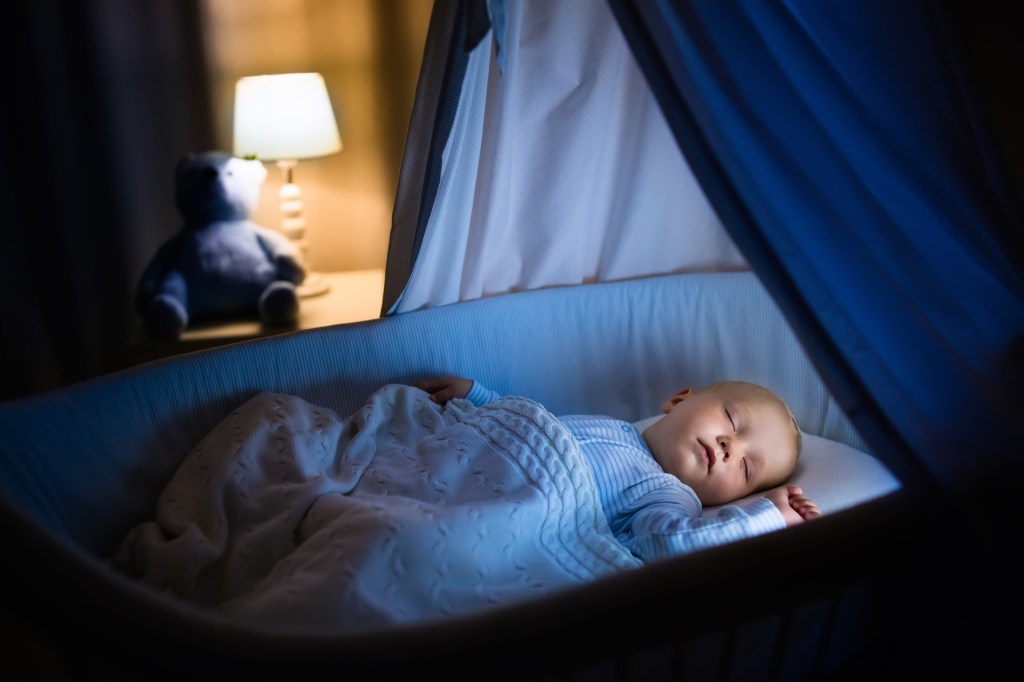 A baby boy sleeps peacefully in a blue bassinet with a canopy at night.