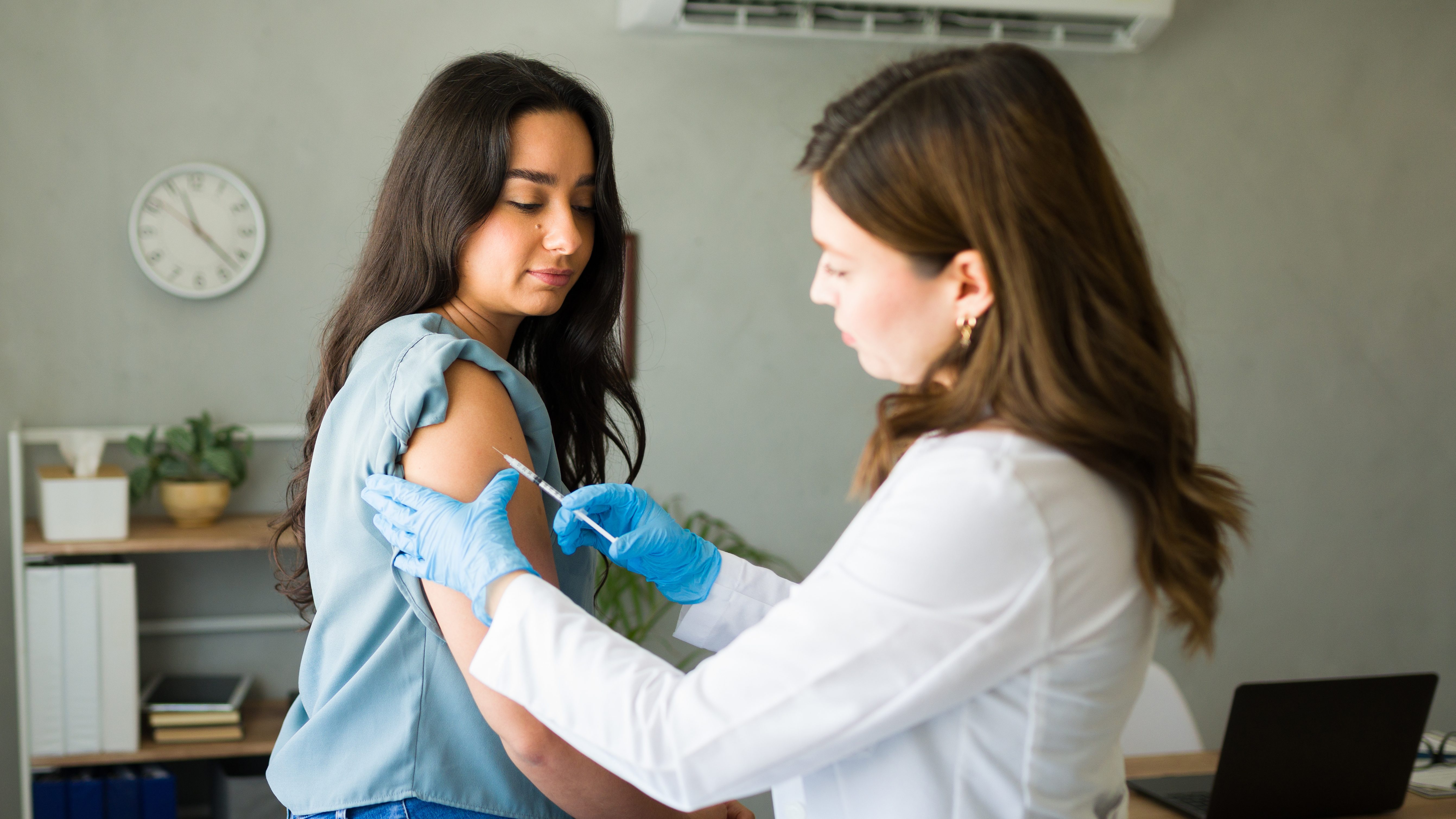 Medical professional wearing gloves administers a vaccine to a young woman in a doctor's office.