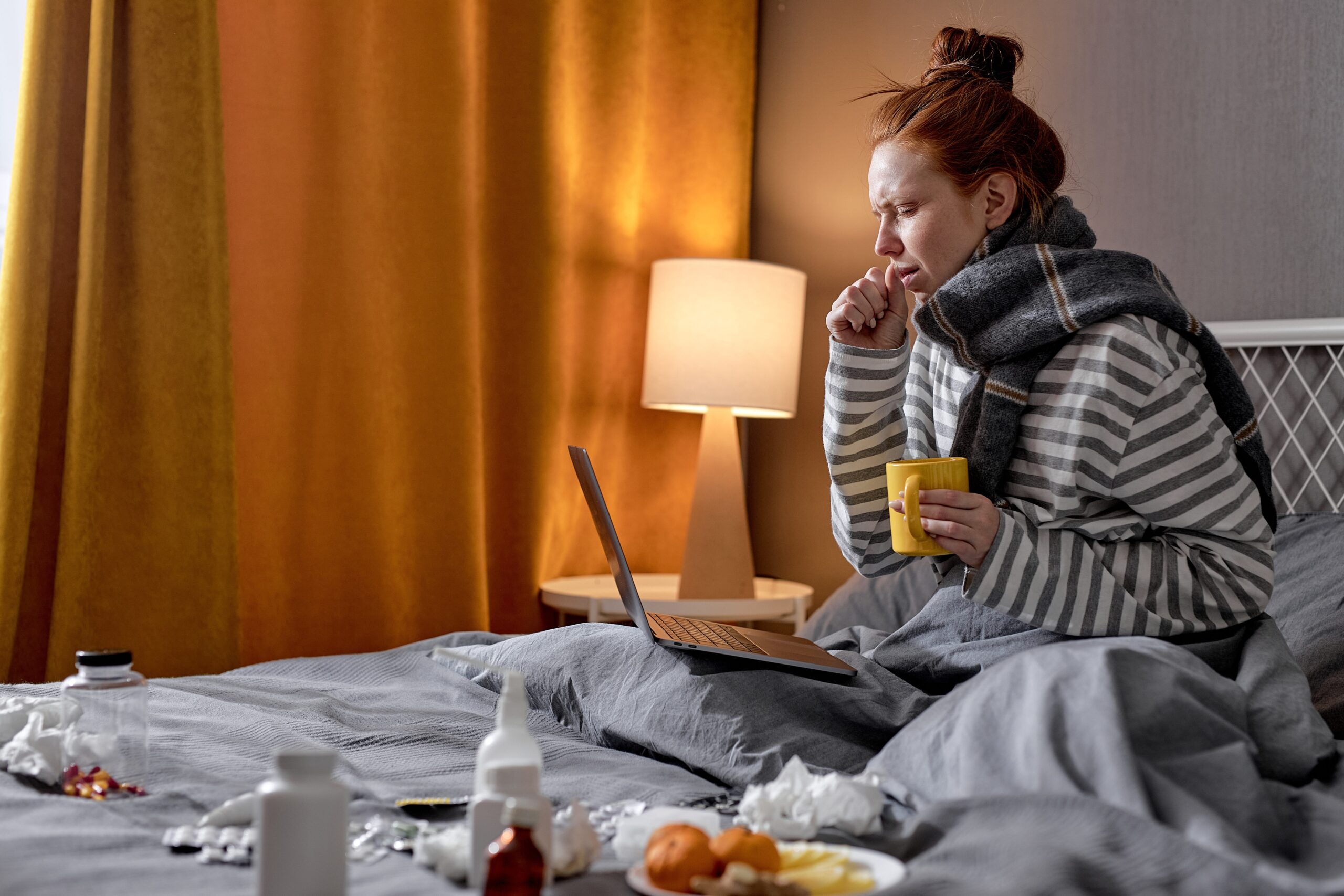 An ill woman with a scarf wrapped around her neck sits in bed, coughing while holding a cup and working on her laptop.