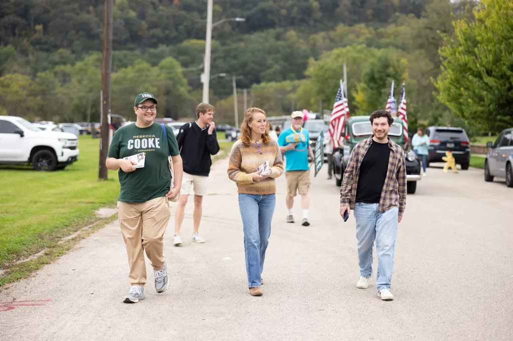 Rebecca Cooke walks down a street with campaign staff and supporters, holding literature as vehicles and American flags line the route behind them.