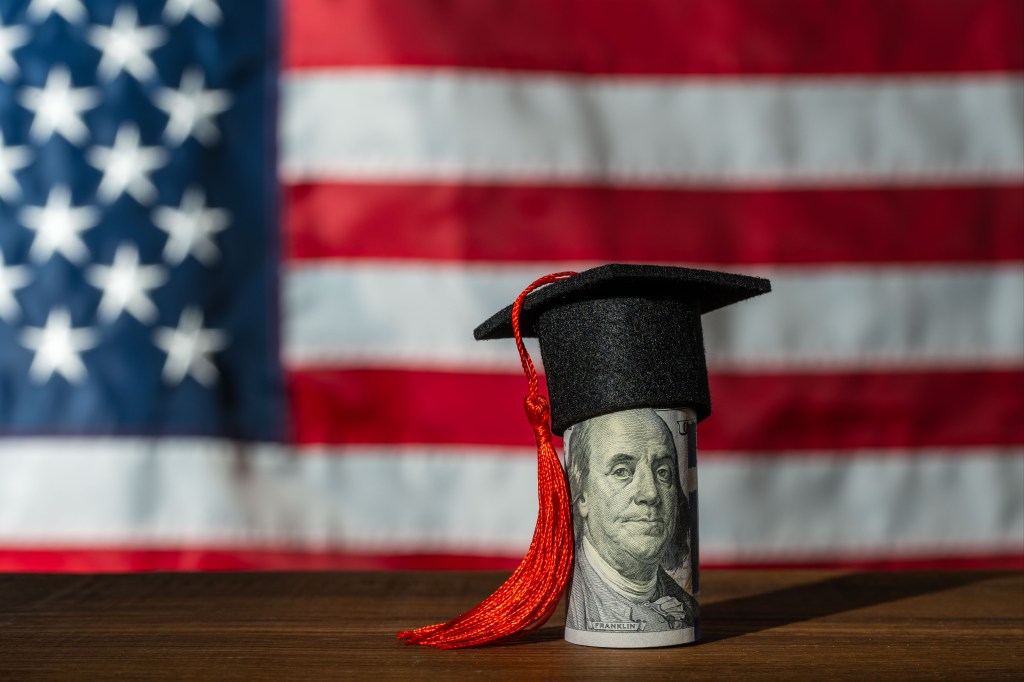 A rolled one-hundred dollar bill with a graduation cap and red tassel in front of a blurred American flag.