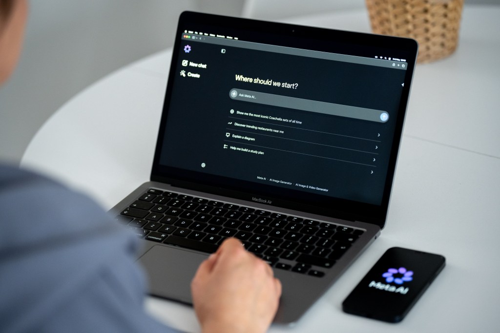 The Meta AI chatbot website displays on a laptop screen, with a smartphone displaying the Meta AI logo lying on the table next to it.