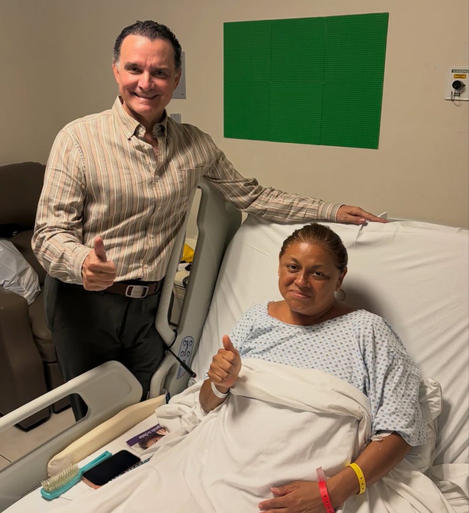 Alma de la Rosa Flores in a hospital bed giving a thumbs up, with her boss, Mario Gamez, standing beside her.