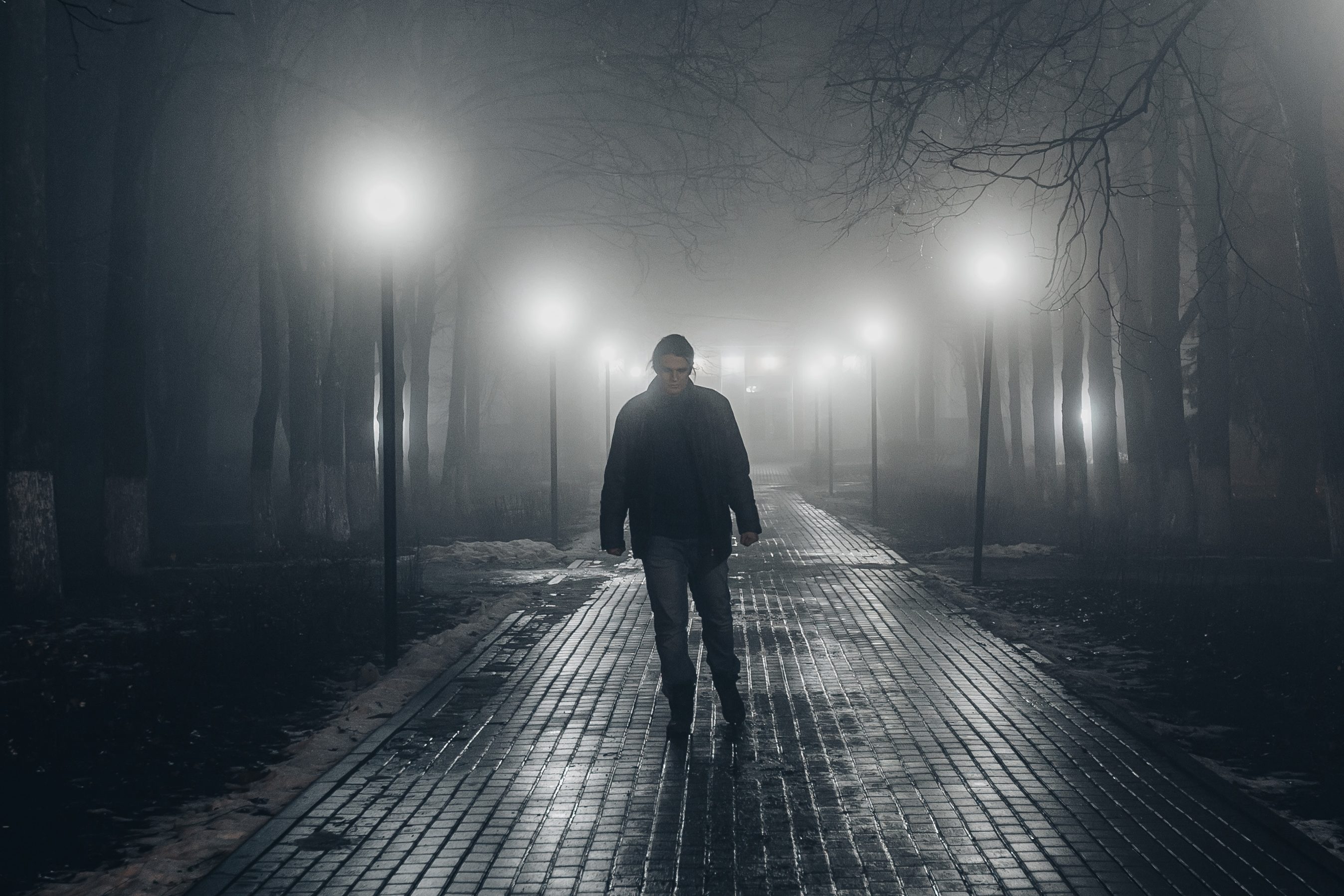 A lone man walking along a dimly lit, foggy park alley at night.