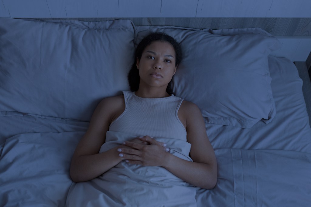 A Black woman lies awake in bed at night with a serious expression, experiencing insomnia.