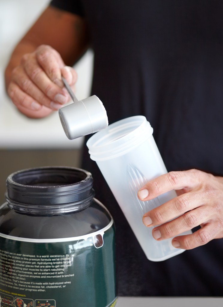 A man preparing a protein shake, pouring white powder from a scoop into a shaker bottle