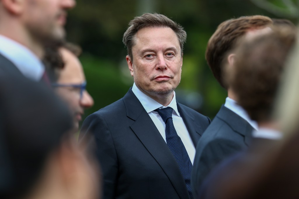 Elon Musk stands in a dark suit and blue tie among other individuals.