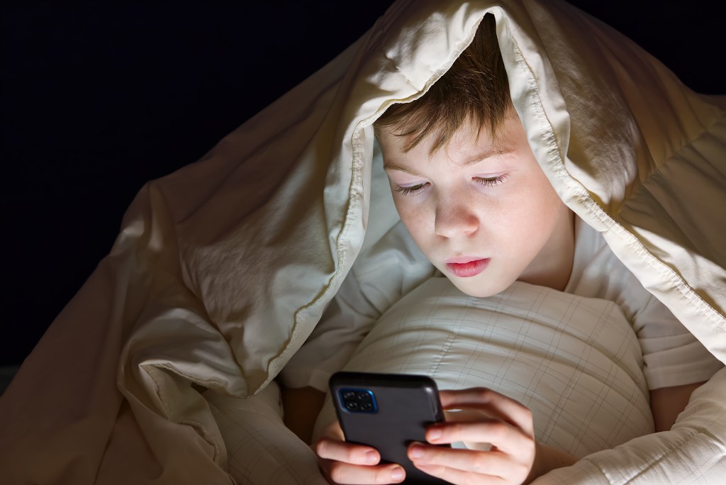 A boy uses a smartphone under a blanket at night.