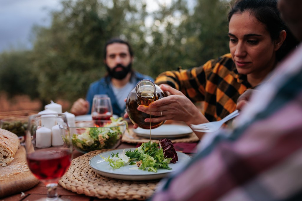 A woman pours olive oil onto a salad during an outdoor meal with friends.