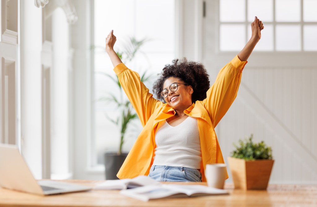 An African American businesswoman rejoices with arms raised in victory while working at a laptop in her home office.