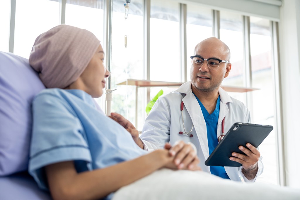 A male doctor holding a tablet reports test results to a female cancer patient in a hospital room.