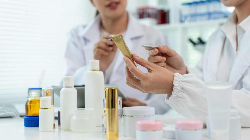 Two female scientists in white lab coats inspect a skincare product tube at a laboratory table surrounded by cosmetic bottles and jars.