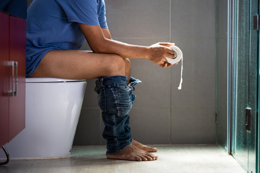 A man holds a roll of toilet paper while sitting on a toilet with his pants down.